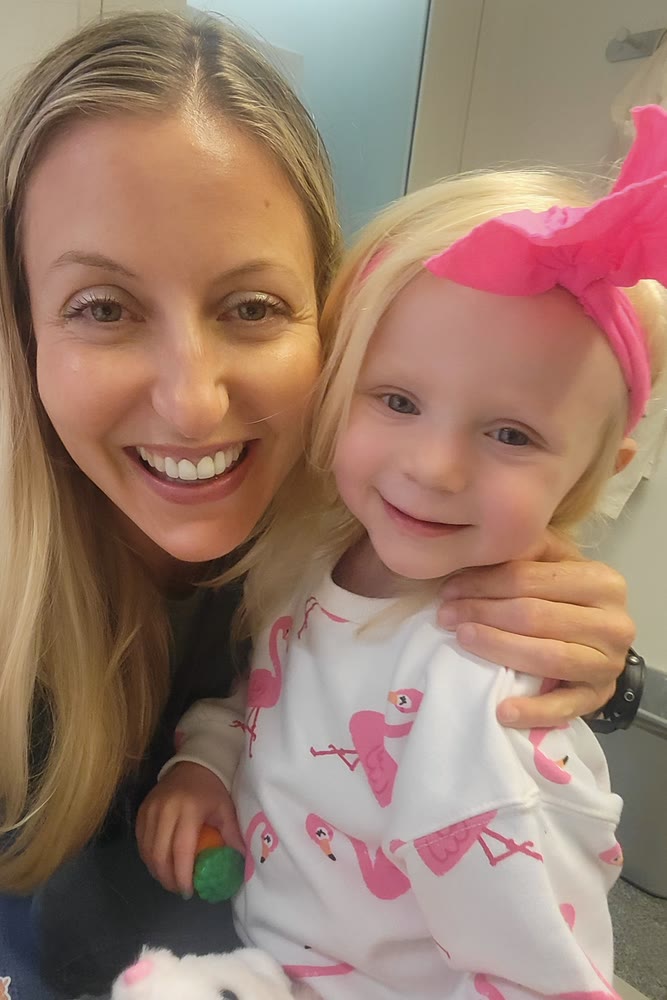 Nicole smiling cheek to cheek with a little girl in a pink bow and flamingo sweatshirt