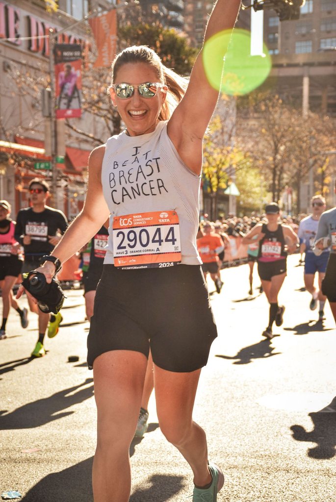 Nicole running the 2024 NYC Marathon with her arm raised, her shirt reading I beat breast cancer